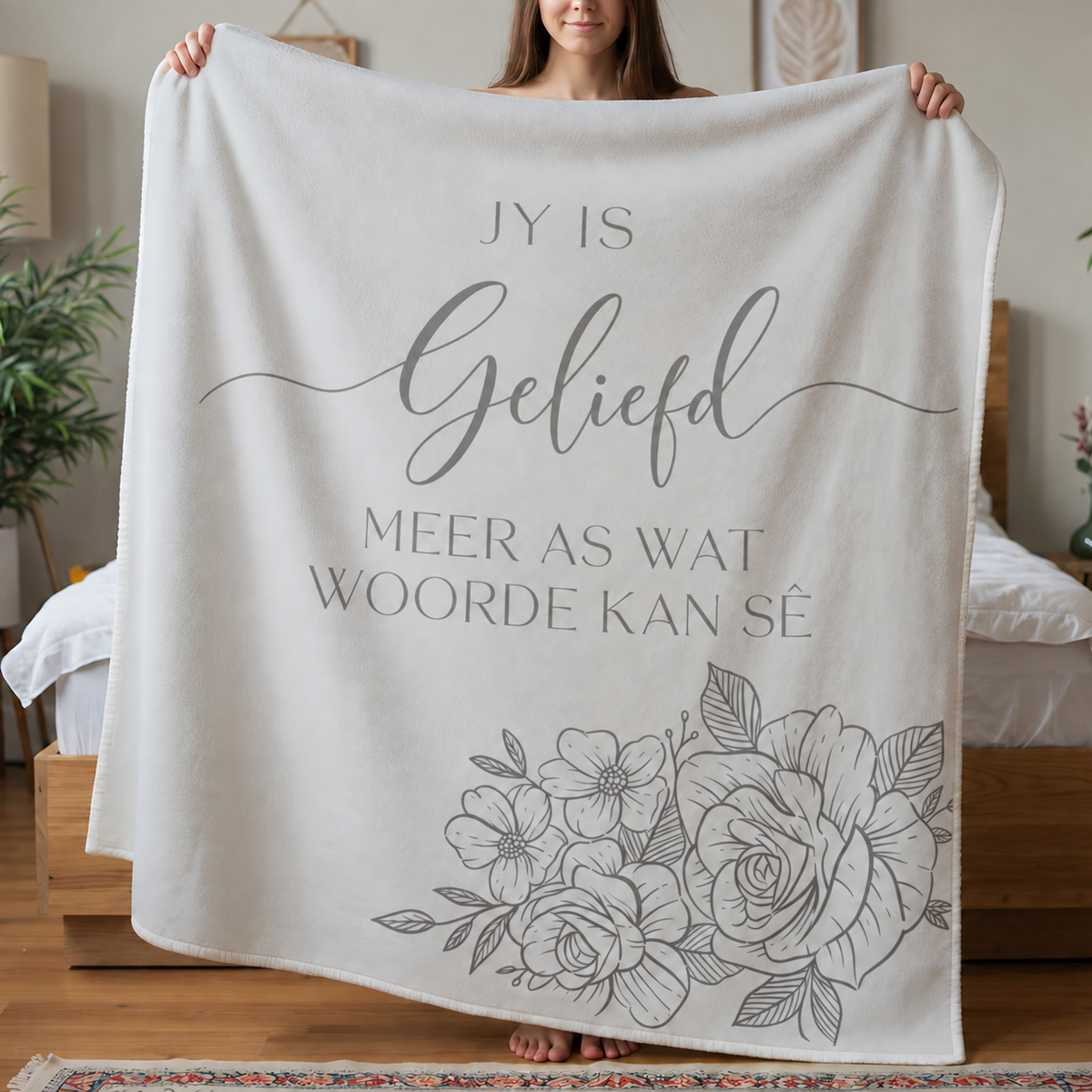 White Afrikaans lightweight fleece blanket with light grey Geliefd wording and floral line art, held by a woman in a bedroom